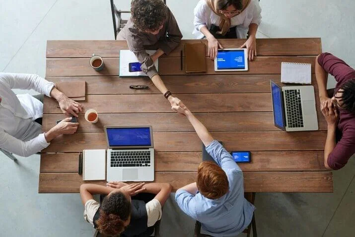 People sit at a wooden table with laptops, notebooks, coffee mugs, and a smartphone. Two individuals across the table from each other shake hands, suggesting a discussion or agreement. The atmosphere appears collaborative and professional.