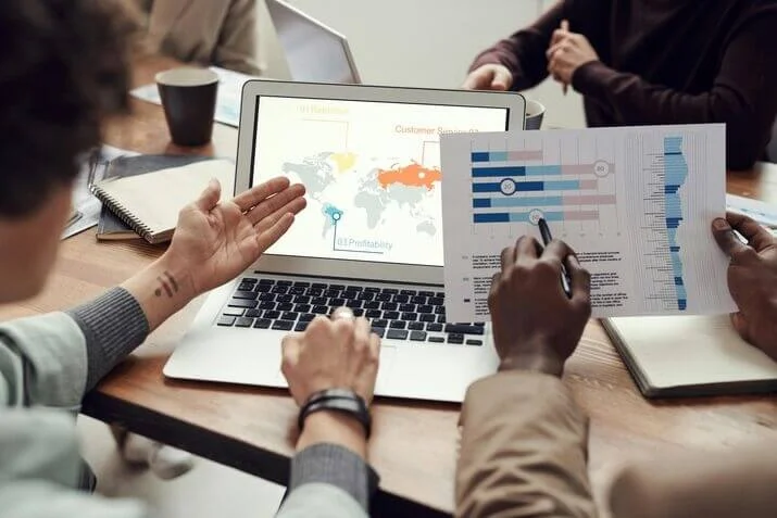People sitting around a table during a meeting, with a laptop displaying a map and chart on the screen. One person is presenting a printed graph, while another is gesturing towards the laptop, discussing IT services. Notebooks, papers, and a cup are scattered on the table.