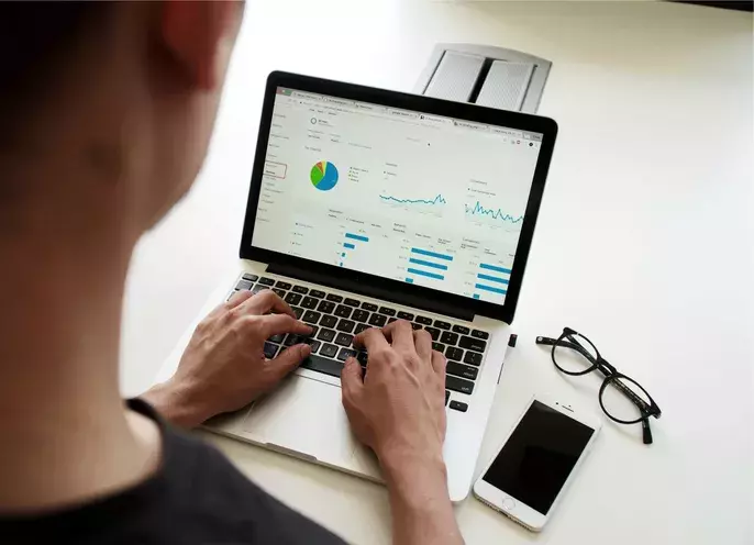 A person is working on a laptop displaying charts and graphs, possibly for an IT audit service. Nearby on the white desk are a pair of eyeglasses and a smartphone. The person appears to be analysing data meticulously.