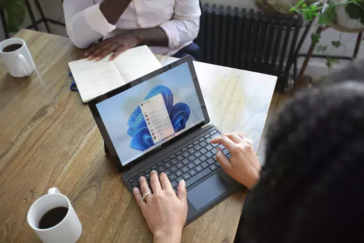 Two people are sitting at a wooden table with notebooks and cups of coffee. One person, possibly from an IT consultancy, is using a tablet with a detachable keyboard displaying a chat app, while the other is writing in a notepad. The scene is set in a well-lit room with plants.