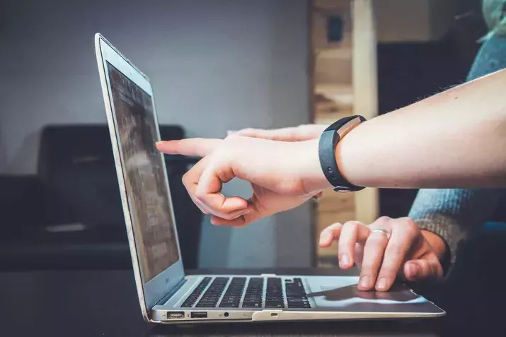 Two hands are seen interacting with a laptop; one hand is pointing at the screen while the other is poised over the keyboard. The scene suggests collaboration or assistance, possibly in a work or study environment, perhaps during security awareness training.