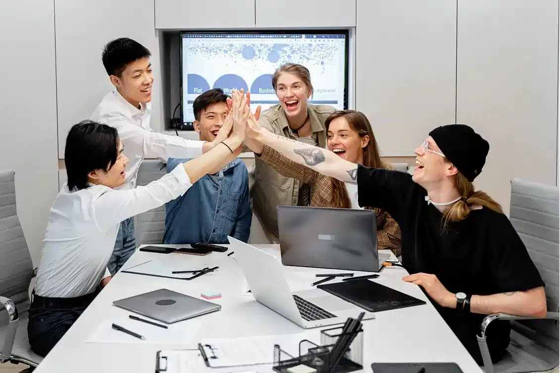 Happy business team celebrating success with a group high-five around a laptop in a modern office, symbolising smooth collaboration and streamlined systems.