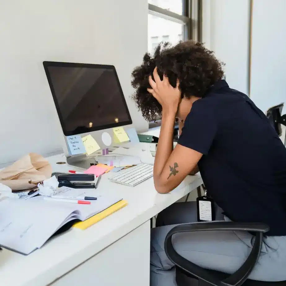 Stressed business owner sitting at a cluttered desk with their head in hands, surrounded by messy papers and an unused computer monitor, representing disorganised systems and tech frustration.